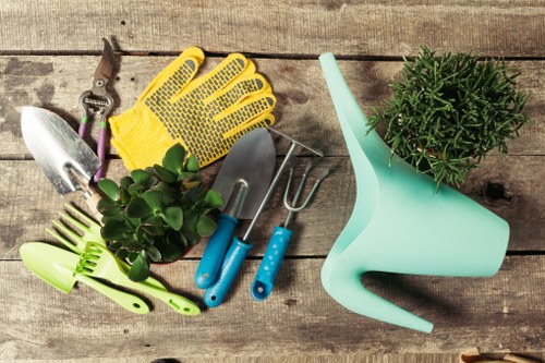 A collection of gardening tools and accessories arranged on a wooden surface, including yellow gardening gloves, small hand trowels with blue handles, a cultivator, and a pair of scissors, alongside a green plant in a black plastic pot and a light blue watering can. The background features aged wooden planks, suggesting an outdoor or garden storage setting. The scene reflects tools typical for gardening and outdoor plant care, with natural textures and colours that highlight a well-organised gardening space suitable for landscaping and maintenance services in Seven Sisters or nearby areas, emphasizing a professional approach to garden care and lawn upkeep.